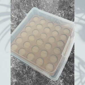 May include: A clear plastic storage box with a wooden insert. The insert has 36 round cutouts for storing small items. The box is closed and the lid is visible.