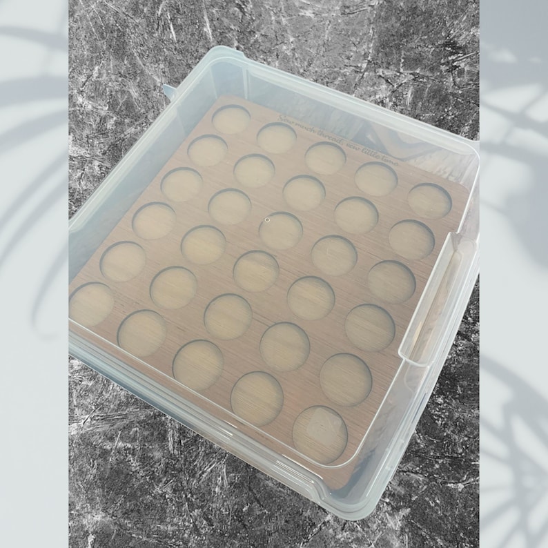 May include: A clear plastic storage box with a wooden insert. The insert has 36 round cutouts for storing small items. The box is closed and the lid is visible.