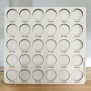 May include: A wooden paint organizer with 36 circular slots for paint pots. Each slot is labeled with a unique code, such as "PF6001" and "PF0423". The organizer is made of light wood and has a natural finish.