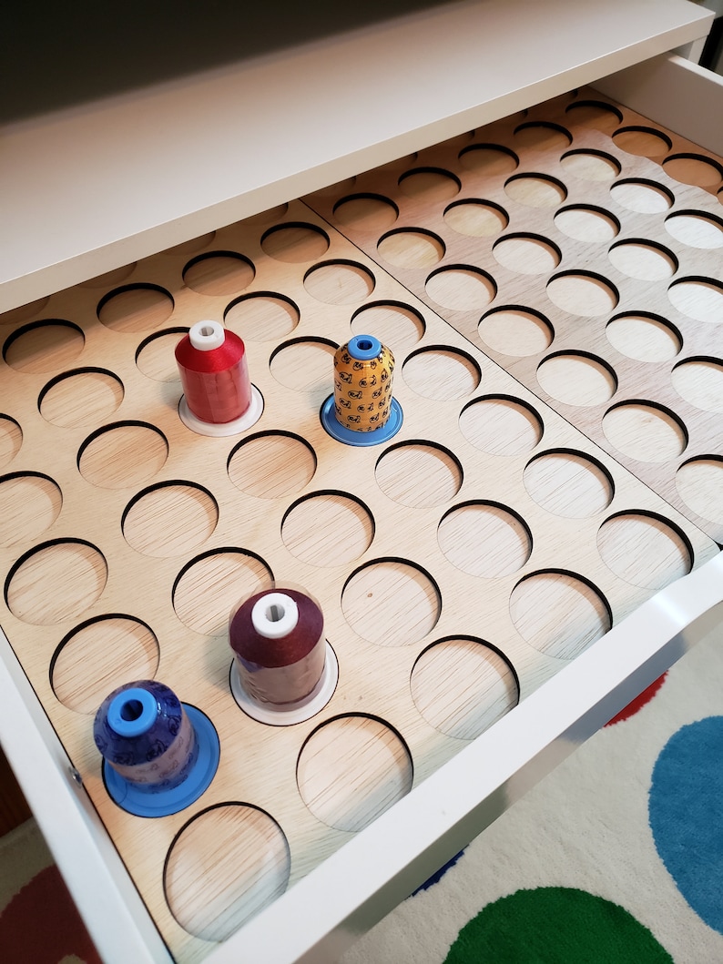 May include: A white drawer with a wooden insert containing multiple circular holes for storing spools of thread. Four spools of thread are visible in the holes, one red, one yellow, one blue, and one maroon.
