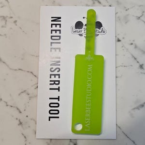 Op de afbeelding: Een limoengroen naaldinrijghulpmiddel met een rechthoekig lichaam en een dunne bovenlip. Het hulpmiddel bevat de tekst LASERBEESTUDIO.COM en een klein bijenicoon. Het is bevestigd op een witte kaart met de tekst NEEDLE INSERT TOOL en een bijenlogo.
