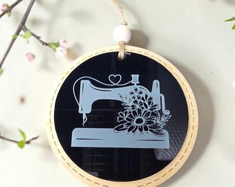 May include: A circular hanging ornament featuring a black acrylic center with a white engraved graphic of a sewing machine adorned with flowers and a heart. The piece is framed by a light-colored wooden border and includes a white bead and twine.
