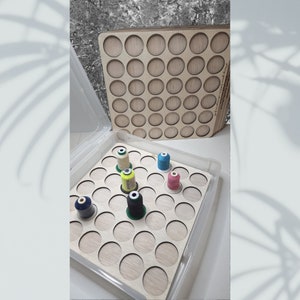 May include: A clear plastic storage box with a wooden insert for organizing thread spools. The insert has 36 circular cutouts for spools. Five spools of thread are visible in the box, in colors green, yellow, blue, pink, and black.