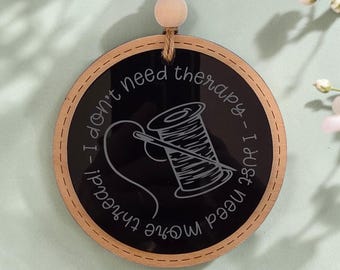 May include: A circular wooden ornament with a black center featuring a white line drawing of a spool of thread and a needle. The text reads, I don't need therapy - I just need more thread! It hangs from a jute twine with a wooden bead.