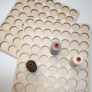 Puede incluir: Dos organizadores de hilo de madera clara con múltiples recortes circulares. Tres carretes de hilo en gris, marrón y crema con centros naranjas se colocan en uno de los organizadores. La veta de la madera es visible.