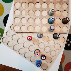 May include: A wooden thread organizer with 72 round slots for storing spools of thread. The organizer is made of light brown wood and has a black border around each slot. There are several spools of thread in various colors, including red, blue, green, and white, in the slots.