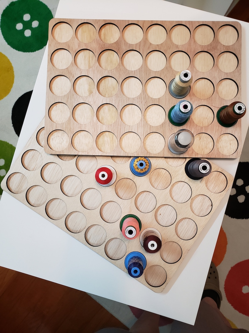 May include: A wooden thread organizer with 72 round slots for storing spools of thread. The organizer is made of light brown wood and has a black border around each slot. There are several spools of thread in various colors, including red, blue, green, and white, in the slots.