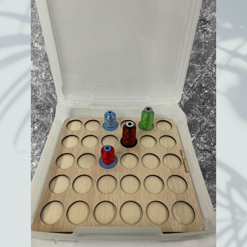 May include: A wooden thread organizer with 25 slots for storing spools of thread. The organizer is in a clear plastic storage box with a lid. There are four spools of thread in the organizer, one red, one green, one blue, and one light blue.
