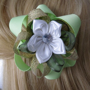 May include: A light green and gold ribbon hair bow with a white satin flower center. The flower has a clear gem in the middle. The bow is set against a background of blonde hair.