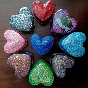 Puede incluir: Una colección de nueve recipientes de resina con forma de corazón dispuestos en círculo. Los corazones presentan varios colores, incluyendo rojo, azul, verde, rosa y morado, con acabados de purpurina. Algunos muestran símbolos blancos.