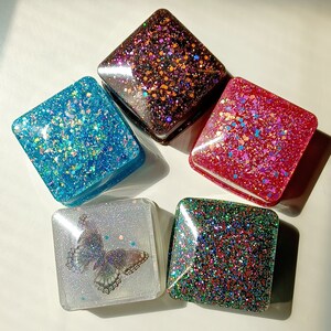May include: Five square resin containers with rounded corners. One is clear with a butterfly graphic, while the others feature dense, multi-colored glitter in black, bright blue, vibrant red, and a dark mix of green, red, and blue sparkles.