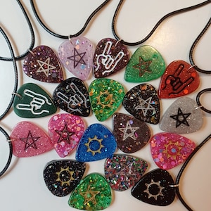 Puede incluir: Una colección de 16 púas de guitarra con varios diseños, incluyendo estrellas, soles y gestos con las manos. Cada púa está hecha de resina transparente con purpurina y un diseño negro o dorado. Las púas están encordadas en cordones negros, creando collares.