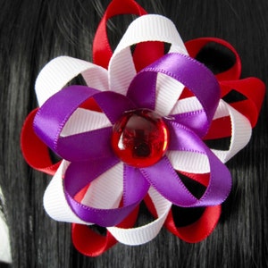 May include: A handmade hair accessory featuring a flower design. The flower is made of red, white, and purple ribbons, with a red gem in the center. The accessory is attached to black hair.
