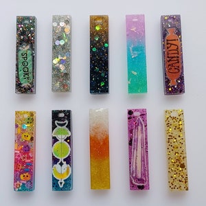 May include: Ten colorful rectangular resin charms with glitter and various designs. The charms include a spooky ghost, a candy cane, a moon and stars, a crystal, and a variety of other designs.