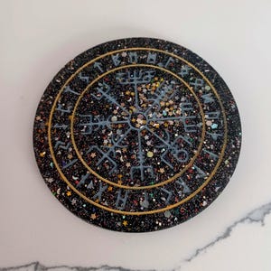 Puede incluir: Posavasos redondo negro con borde dorado y diseño celestial. El posavasos presenta un símbolo central rodeado de runas e incrustado con purpurina y detalles en forma de estrella. El diseño es en tonos azules y dorados.
