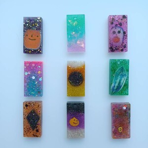 May include: Nine rectangular resin pendants with glitter and various designs. The pendants feature images of a pumpkin, a skull, a crystal, and other Halloween-themed elements. The pendants are in various colors, including purple, pink, and orange.