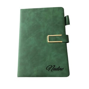 Könnte beinhalten: Ein grünes, ledergebundenes Notizbuch mit einem goldfarbenen Verschluss und dem Namen "Nadine" in Schreibschrift. Das Notizbuch hat eine strukturierte Oberfläche und einen passenden grünen Laschenverschluss.