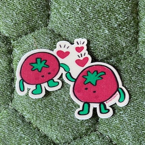 May include: Two red tomato stickers with green stems and feet, holding hands, with a white heart-shaped figure and red hearts above them. The stickers are on a textured green background.