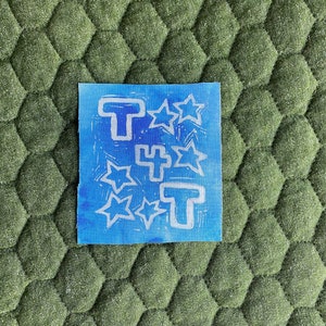 Puede incluir: Una tela azul con una impresión de tinta blanca de tres letras T estilizadas y cinco estrellas.