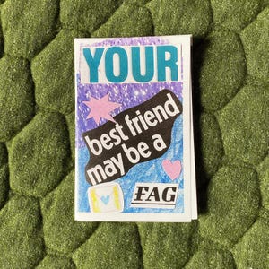 May include: A small, folded paper with the words "YOUR best friend may be a FAG" in a mix of fonts and colors. The background is a mix of purple and blue with a pink star and heart. The paper is on a green, textured surface.