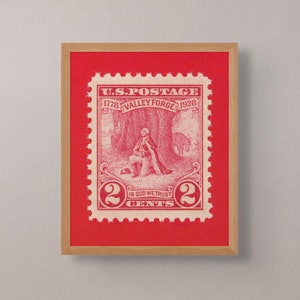 May include: Framed vintage U.S. postage stamp, featuring a red and white design with the text "U.S. POSTAGE" and "VALLEY FORGE 1778 1928". The stamp depicts a historical scene and the text "IN GOD WE TRUST". The frame is wood.