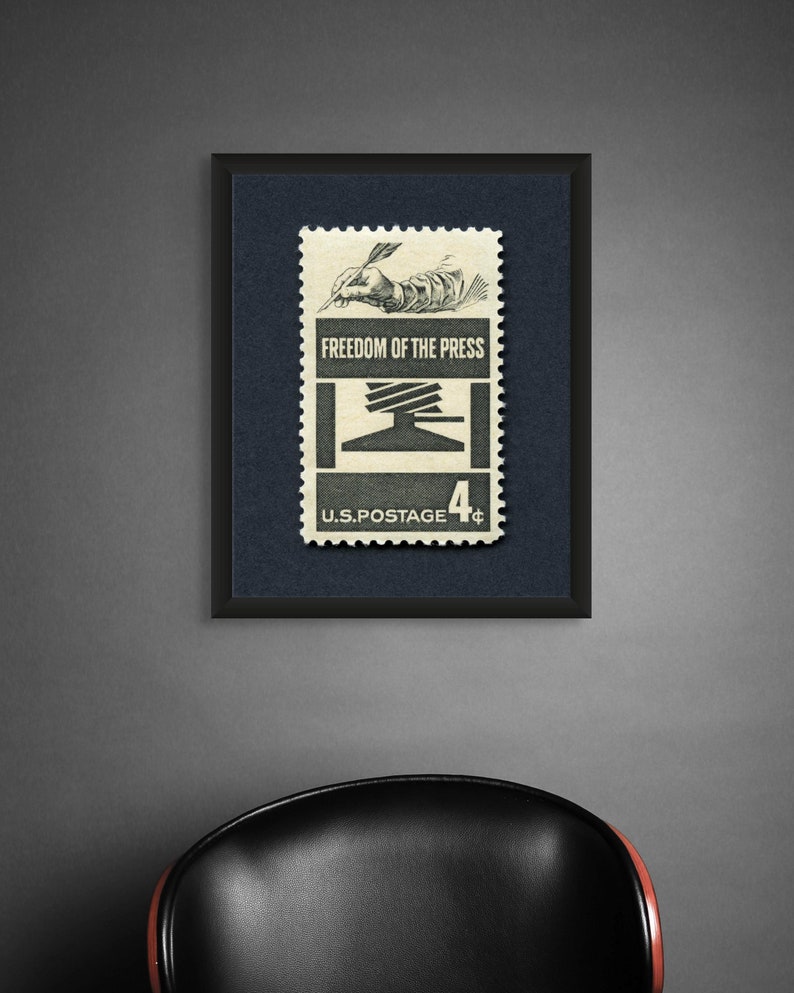 May include: A framed vintage US postage stamp with a black frame. The stamp features a black and white illustration of a hand writing with a quill pen. The stamp reads "Freedom of the Press" and "U.S. Postage 4&cent;".