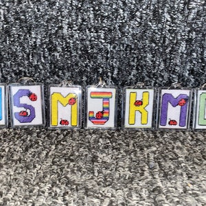 May include: A set of seven colourful embroidered keychains with the letters H, S, M, J, K, M, and D. Each keychain features a small red ladybug.