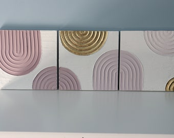 May include: Three square canvases with a textured, abstract design. The canvases feature a combination of pink and gold paint, creating a modern and minimalist look.