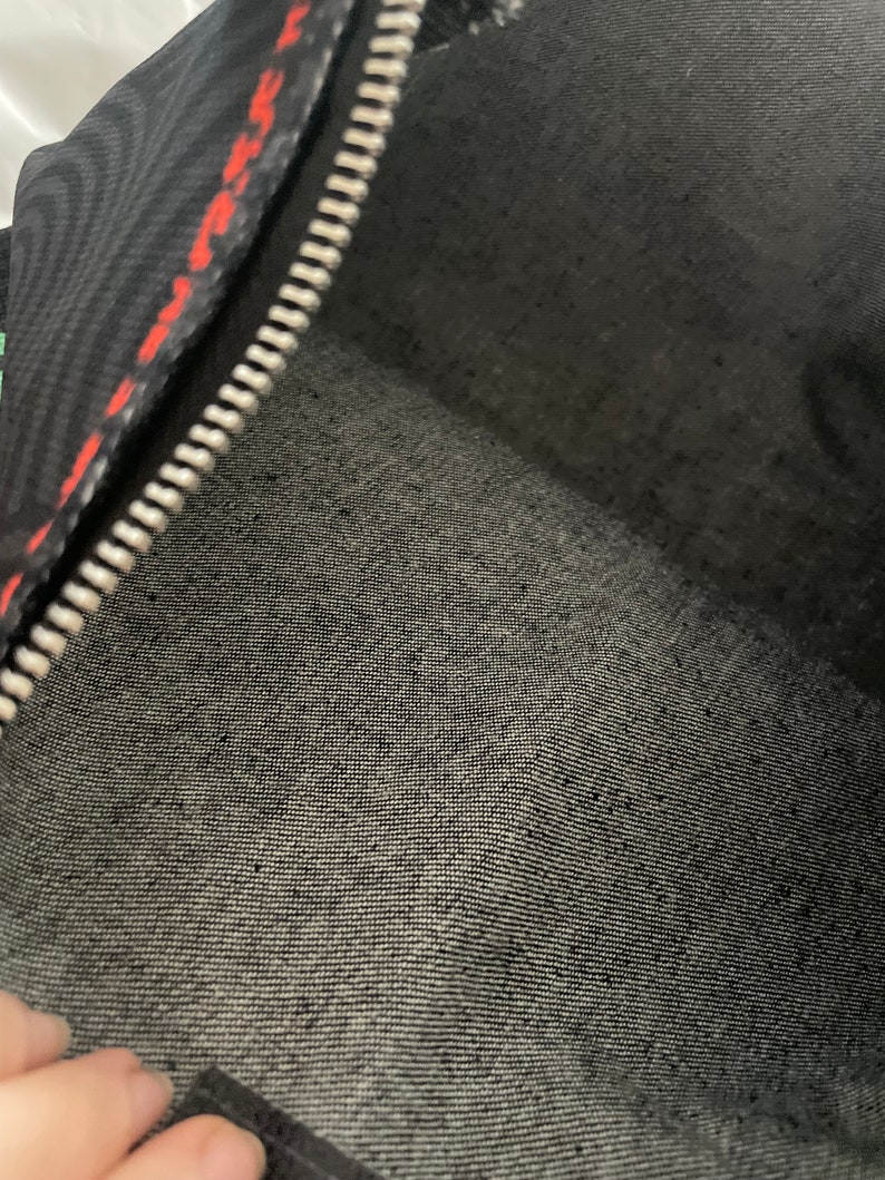 May include: Black zippered pouch with a red and black patterned lining. The pouch is made of a soft, textured fabric.