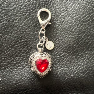 May include: Silver heart-shaped cremation keepsake with a red crystal and a silver charm with the letter "Z" on a silver chain.