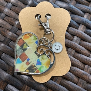 May include: A silver heart-shaped keychain with a colorful collage inside. The keychain has a silver charm with the letter "G" on it.