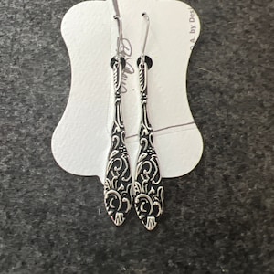 May include: A pair of silver dangle earrings with a black filigree design. The earrings are on a white card with the text "A. by Desi" on it.
