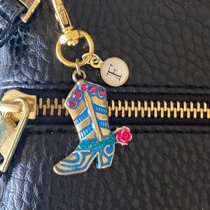 May include: A gold charm with a blue and gold boot with pink and green accents and a pink rose. The charm is attached to a gold chain and a gold circle with the letter "E" on it.