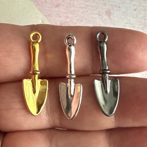 May include: Three small metal charms shaped like shovels. The shovels are gold, silver, and black.