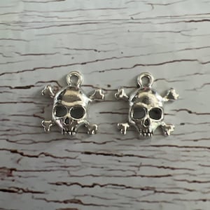 K&ouml;nnte beinhalten: Zwei silberne Totenkopf- und Knochen-Anh&auml;nger. Die Totenk&ouml;pfe sind detailliert gearbeitet und an einer Schlaufe zum Aufh&auml;ngen befestigt.