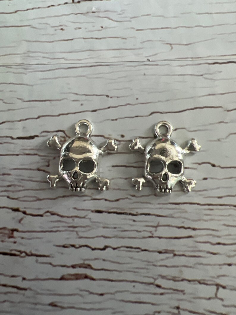 K&ouml;nnte beinhalten: Zwei silberne Totenkopf- und Knochen-Anh&auml;nger. Die Totenk&ouml;pfe sind detailliert gearbeitet und an einer Schlaufe zum Aufh&auml;ngen befestigt.