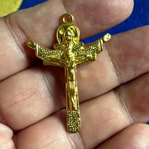 May include: A gold-toned metal crucifix pendant featuring a textured surface on the cross arms and base. The design includes a central figure of Jesus on the cross with a smaller figure positioned above the head, all topped by a circular loop.