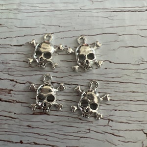K&ouml;nnte beinhalten: Vier silberne Totenkopf- und Knochen-Anh&auml;nger. Jeder Anh&auml;nger zeigt einen detaillierten Totenkopf mit Knochen dahinter. Die Anh&auml;nger haben eine Schlaufe oben zum Anbringen an Schmuck oder anderen Bastelarbeiten.