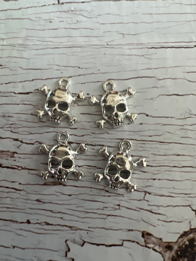 K&ouml;nnte beinhalten: Vier silberne Totenkopf- und Knochen-Anh&auml;nger. Jeder Anh&auml;nger zeigt einen detaillierten Totenkopf mit Knochen dahinter. Die Anh&auml;nger haben eine Schlaufe oben zum Anbringen an Schmuck oder anderen Bastelarbeiten.