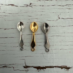 May include: Three small spoons, one silver, one gold, and one nickel, are arranged in a row on a white wooden surface.