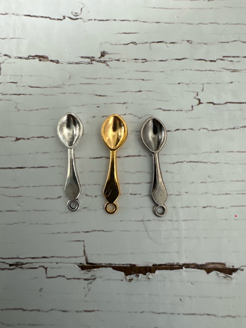 May include: Three small spoons, one silver, one gold, and one nickel, are arranged in a row on a white wooden surface.