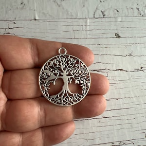 May include: Silver-toned metal pendant featuring a detailed tree of life design. The pendant is circular and has a delicate, intricate pattern.