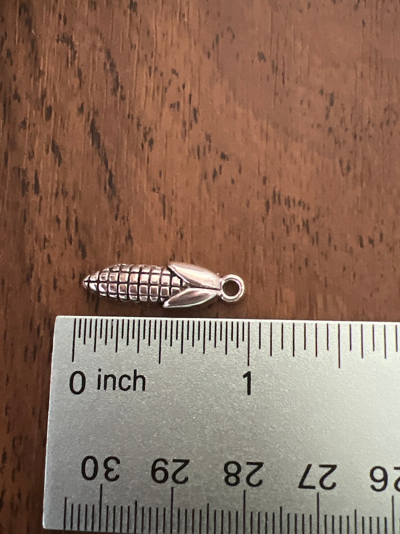 May include: Silver charm shaped like an ear of corn, measuring about 1 inch long. The charm has a loop at the top for attaching to a bracelet or necklace.