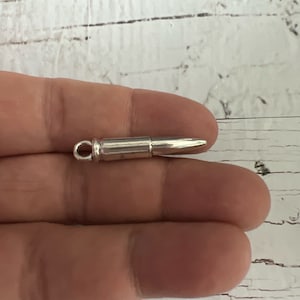 May include: A silver bullet charm pendant. The charm is shaped like a bullet and has a small loop at the top for attaching to a chain or bracelet.