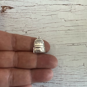 May include: A silver charm in the shape of a backpack. The charm is detailed with a zipper and straps.