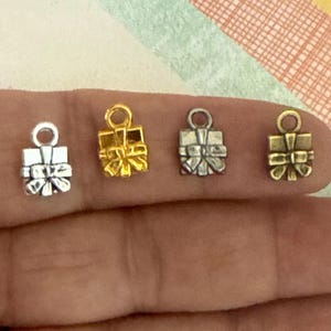 May include: Four silver and gold gift box charms with a bow on top. The charms are on a hand.