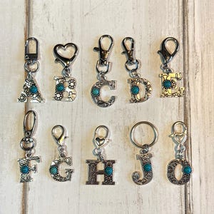 May include: A collection of silver-toned initial keychains with turquoise-colored accents. Each letter, from A to O, features a decorative design and a lobster clasp for easy attachment. The keychains are displayed on a light wood background.