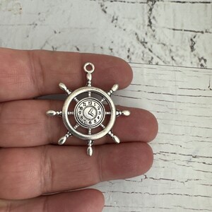 May include: Silver-toned metal charm shaped like a ship's wheel with a clock face in the center. The clock face has Roman numerals and a heart-shaped hand.