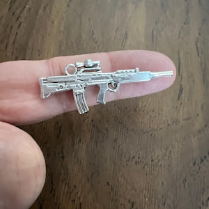 May include: Silver charm of a rifle with a scope. The charm is small and detailed.