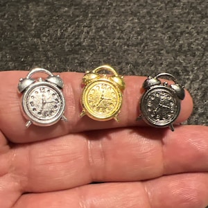 May include: Three miniature alarm clocks in silver, gold, and black metal. Each clock has a round face with black Roman numeral hour markers and black hands.